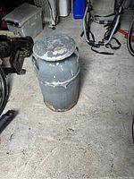 Photo of an antique metal milk can with gray paint, showing chipping and rust on the lid and body, placed on a concrete floor among bicycles and other items.
