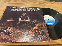 Vinyl LP record and cover of Songs From The Wood by Jethro Tull with rustic forest campfire cover art.