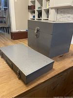 Two vintage metal boxes on a wooden table, one larger textured gray box with latch lock and keys, one smaller flat silver box with handle and latches.
