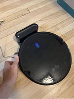 Top view of Eufy RoboVac 11S black circular robot vacuum with blue power button illuminated on wooden floor near charging dock.