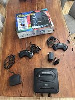 Sega Genesis Model 2 console with two controllers, power adapter, Sega Menacer adapter, and Aura Interactor with original box on wooden table.