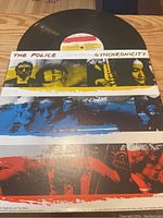 Top view image of the black vinyl record partially removed from its sleeve. The sleeve has colorful band member portraits in yellow, blue, and red with the text 'The Police Synchronicity'. The label on the record is visible with the album's title.