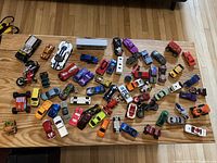 Overhead photo showing approximately 40 toy cars, trucks, motorcycles, and trailers in various colors arranged on a flat wooden floor.