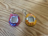 Top front view of pink and yellow Tamagotchi toys showing screen, buttons and keychains.