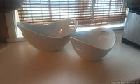 Two large white ceramic serving bowls with handles. The bowls have a rounded shape and smooth glazed finish. Placed on a counter near window blinds.