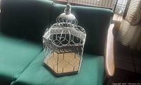 Large white metal birdcage with hexagonal shape sitting on a green sofa showing the entire cage and its size.