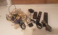 Lot contents showing various tangled electronic cables, power adapters, and three remote controls lying on a white surface.