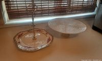 Two cake trays on a flat surface near a window; left tray features floral antique plate on stand with gold handle, right tray is white ceramic pedestal cake stand with spiral design