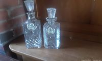 Two crystal liquor decanters on a wooden shelf showing the full view with stoppers and porcelain tags.