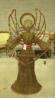 Full view of the large woven angel figure showing the entire structure with wings spread and halo above the head, made from natural twigs with string lights wrapped around.