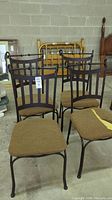 Set of four black metal frame chairs with brown foam seat cushions, one cushion torn revealing foam. Chairs show wear marks on tops.