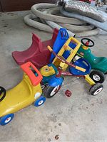 Shows four riding toys grouped: yellow Little Tikes push car, red and blue Radio Flyer tricycle, green and yellow Little Tikes ride-on lawn mower, and red resin see saw