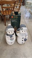 Two snow people figurines side by side with the fiber optic tree box behind them on garage floor. One snow person has a damaged top of head.