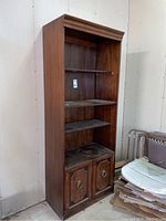 Front view of tall wood display cabinet showing four shelves and lower two-door storage with ring handles. Medium brown wood finish, heavily used with dust.