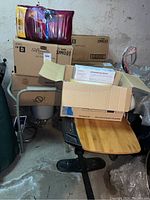 Photo showing stacked boxes of Silhouette adult diapers, cardboard box with poly gloves, metal frame commode chair, and wooden top meal tray on concrete floor in basement.