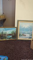 Two framed paintings leaning against a wall on a carpet, one larger seascape in light green/gold frame, one smaller riverside scene with wood frame showing paint cracking on canvas.