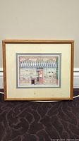 Framed print of a bakery storefront with blue and white striped awning, signed print, wooden frame natural finish