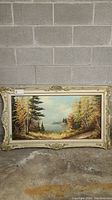 Full view of the framed landscape painting showing autumn trees along a water body under a cloudy sky, set in an ornate cream wood frame with evident damage.