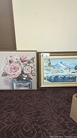 Two paintings side by side on floor; left painting floral with pink roses, right painting winter landscape with snow and cabins. Both framed.