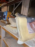 Stack of four unopened memory foam contour pillows with visible packaging on basement shelf along with other items behind