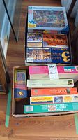 Top down view of boxed puzzles stacked on hardwood floor, including Ravensburger, Peeluxe Deluxe, and Kodacolor boxes.