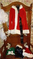 Full view of Santa suit jacket hanging on wooden chair with black belt. White synthetic beard hanging on the left side of the chair. Red pants and black boot covers with white fur trim along with Santa hat and green Christmas-themed bag sitting on seat.