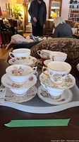 Ten assorted porcelain teacups and matching saucers displayed on trays, various floral and gold designs, one footed cup visible
