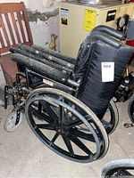 Side view of the folded wheelchair showing black padded seat and backrest, large rear wheels and small front caster wheel, dusty condition.