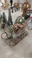 Wire and wicker sleigh holding decorative wrapped presents, with Christmas light garland, shown in warehouse environment with other furniture and decorations.