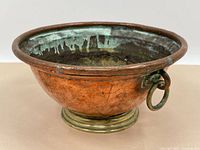 Front angle view showing the copper bowl with green oxidation inside and around the rim and the attached round brass handles on each side.