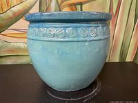 Blue ceramic pottery planter with decorative circular band near rim, placed on a black surface with colorful background.