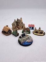 Overview of seven ceramic model buildings and houses arranged in a group, varied shapes and colors. Includes church model, stone cottages, and a base model with water and boat.