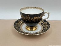 Tea cup and saucer set with black enamel and gold leaf floral detailing on a white background, ornate handle with gold accents, shown on white surface.