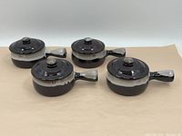Four dark glazed stoneware soup bowls with speckled light gray handles and lids, arranged in two rows on a tan surface.