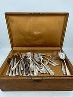 Wooden box opened displaying assorted silverplate knives, forks, and spoons inside a khaki-colored fabric lining with Daly Lavoisy branding.