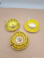 Photo showing three vintage tea cup and saucer sets with varying yellow and gold designs including floral and scrollwork patterns.