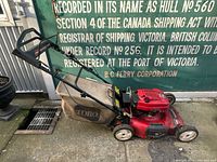Side view of the red Toro GTS 6.75 HP walk behind lawn mower positioned next to a wall with signage. Grass collection bag attached and handle visible.