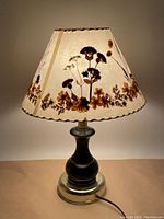 Photo of the vintage table lamp lit, showing the floral pressed flower design on the beige fabric shade and the dark and brass-toned base.