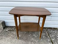 View of the entire wood end table outdoors showing angled rectangular top and four tapered legs with brass caps.