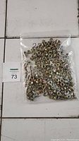 Plastic bag containing approximately 250 hex cap serrated flange nuts placed on a tiled surface with maxsold lot card beside it.
