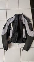 Full view of black and grey Daymak biker motorcycle jacket laid flat on white tiled floor, showing front zipper and arm panels.