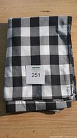 Folded black and white checkered buffalo plaid tablecloth with label showing lot number 251, placed on wooden surface.