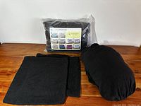 Two black fabric sofa covers displayed on a wooden surface; one folded and one rolled, alongside a packaged sofa cover with label showing various sofa prints.