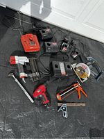 Overview of the assortment of Ryobi hand tools including chargers, drill, impact driver, ratchet wrench, caulking gun, circular saw, and combination square placed on a black cloth.