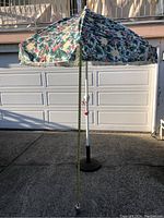 Outdoor floral umbrella fully opened with a measuring tape showing height from ground to top canopy of about 74 inches, white pole, black weighted base stand in concrete driveway in front of garage.