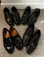 Four pairs of men's dress shoes arranged on the floor, showing two black slip-on pairs, one brown lace-up pair, and one pair of black tassel loafers.