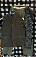 Full view of olive vest, pants, and scarf hanging together against black and white checkered background