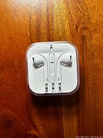 Front view showing Apple EarPods wired earphones neatly packaged inside a square clear plastic case with visible inline remote and mic.