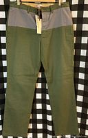 Front view of men's pants showing color-block design in olive green and gray, labeled size 34x32 with tags attached.