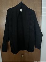 Front view of new black ribbed knit women's sweater showing open front and shawl collar hanging on wooden door.